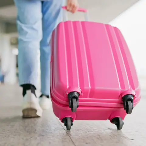 Getty Images A young woman wearing light jeans and white trainers pulling a bright pink suitcase in an airport
