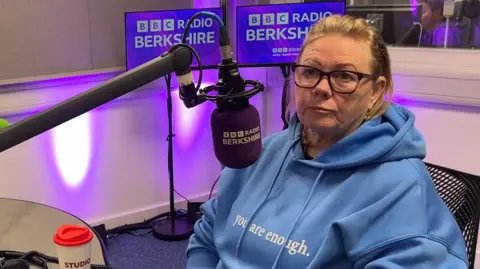 Helena Tym is sat in BBC Radio Berkshire's studio in front of a branded purple microphone. She is wearing a blue hooded top which says "you are enough". 