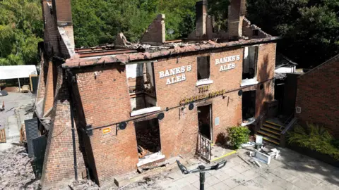 A burnt-out shell of a red brick two-storey former pub building, with roof and windows missing. The Crooked House can just be seen in lettering above the former door, with Banks's Ales stencilled on the outer wall above. Three window cavities can be seen on the top level, and two plus a door cavity on the bottom. Rubble can be seen inside the building, spilling out onto a patio. There are trees behind the building and a concrete patio in front.