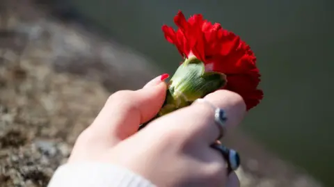 A woman holds a red carnation in her hand at the National Covid Wall of Remembrance in London. You can see rings on her fingers and a red manicure.