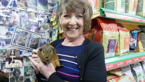 Family Handout Barbara is holding a guinea pig in her pet shop with bags of food on shelves behind her. She is smiling.