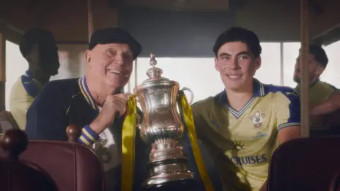 Southampton Football Club Mickey Channon, an older man wearing a flat cap, sits in a bus with a vintage interior while holding onto one side of the FA Cup trophy. The other side is held by Jay Robinson, who's sat on the opposite side of the aisle. Both men are smiling. Behind them, on either side of the bus, are Joshua Quarshie and Ryan Manning, both obscured slightly by shadow. 