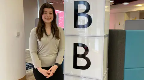 A woman in a grey top and black trousers with long brown hair smiling and holding her hands together