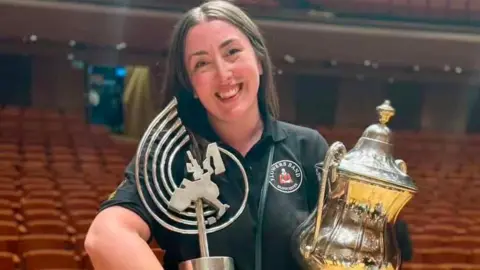 A woman called Stephanie Binns holds two silver trophies in a concert hall she won with The Flowers Band. She is smiling. She is wearing a black polo shirt with her band's logo on it. Binns has long brown straight hair.