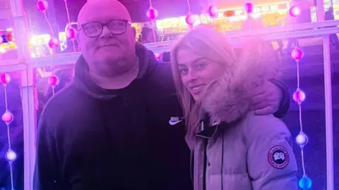 Stephen Brady, who has a bald head, black framed glasses and is wearing a black zip up Nike branded hoodie, stands in front of a light installation with his arm around the shoulders of his daughter Ellie-May, who has long blonde hair and is wearing a beige coat with a large fur hood.