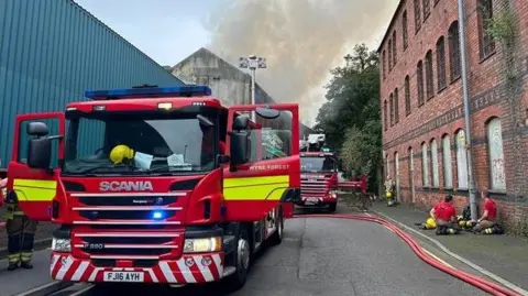 Hereford and Worcester Fire and Rescue Service Two fire engines on Park Lane, Kidderminster. Smoke can be seen drifting in the distance and fire fighters are sat on the floor next to their equipment.