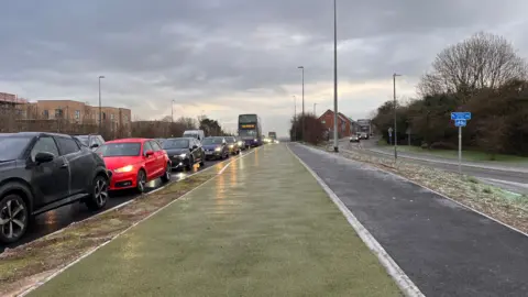 A green cycle lane next to a grey cycle path, pictured facing the incoming traffic, in a grey icy morning.