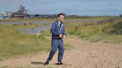 Selebaran keluarga Declan berlarian di dekat pinggir laut di Skegness. Dia mengenakan baju olahraga biru dan sepatu kets hitam. Sebuah bianglala dan pasar malam di latar belakang