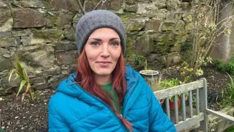 A woman with red hair wearing a blue coat and grey wool hat sat on a garden bench