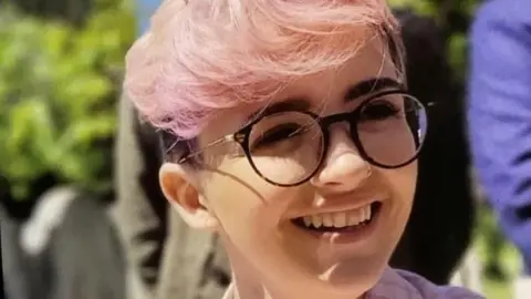 Family Handout A woman with short pink hair wearing black glasses and smiling.