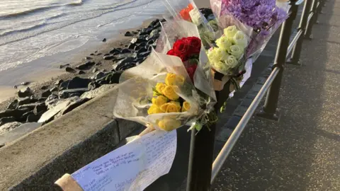 Luke Deal/BBC Floral tributes and cards on a barrier next to rocks and the sea