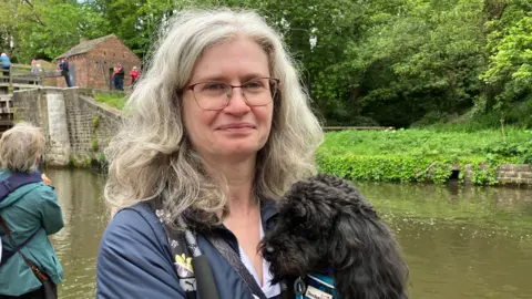 Olivia Richwald/BBC A woman stands in front of a canal. She wears glasses and a blue coat and holds a medium-sized black dog in her arms.