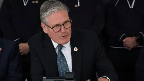 Sir Keir Starmer smiles while speaking on the phone to President Trump from a car factory in the West Midlands.  He is wearing a dark colour suit with a blue patterned tie and has a poppy lapel pin to signify the 80th anniversary of VE Day. 