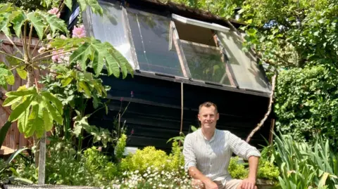 Nick Fry A black shed with glass panels pointing towards the sky. A man is sat in front of it, with his hands on his lap.