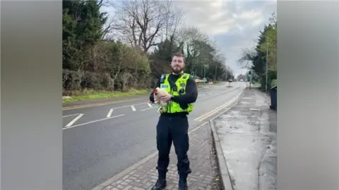 Cambridgeshire Constabulary A police officer stands to the side of an empty road holding a white chicken. 