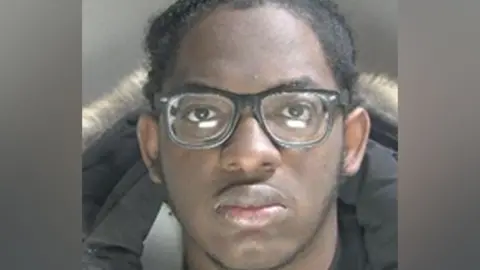 A police custody image of Sakeen Gordon, wearing black-rimmed glasses. He is looking straight at the camera. He is wearing a black coat with a fur trim. He is not smiling. 