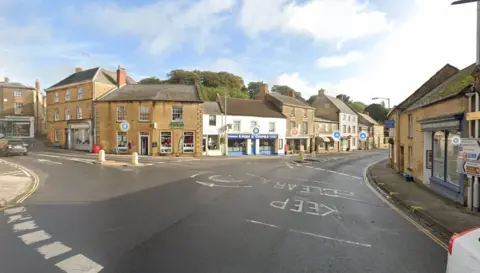 A road with market square in Crewkerne. It has a mini-roundabout and shops in the background.