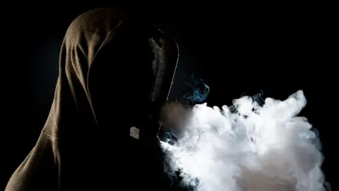 An anonymous man in a black hoodie smokes a vape and exhales.