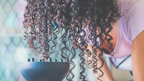 Getty Images A person uses a hairdryer fitted with a large diffuser attachment to dry curly hair. The person's head is bent forward so that long, dark ringlets hang down towards the diffuser.
