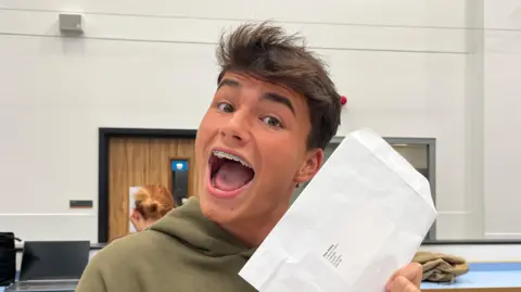 Finn is holding the white envelope aloft in both hands and has his mouth open in a wide smile for the camera. He has short brown hair which is spiked up at the front and is wearing a green hoodie.