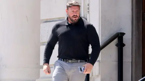 Karl coming out of court. He has short brown hair and a groomed moustache and beard. He has a black top on and grey jeans. he holds his phone in his left hand.