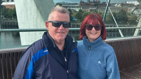 Man standing on the left with grey short hair, wearing sunglasses, a zip up blue and navy coat with a zip up navy hoodie underneath. Woman on the right has short red hair, wearing large sunglasses and a half zip fluffy blue fleece.