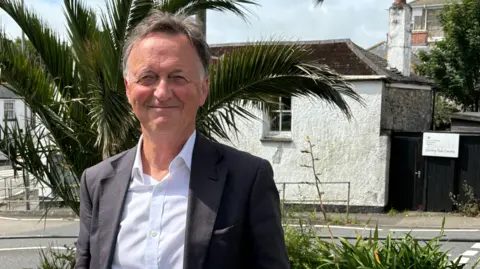 A man with greying hair wearing a blue shirt and dark jacket standing to the left of the picture with the Penzance driving test centre behind him.