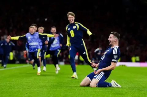 PA Media Scotland's Kenny MacLean is on his knees on the Hampden pitch as he celebrates scoring his goal against Denmark. Scotland substitutes run towards him to congratulate him.