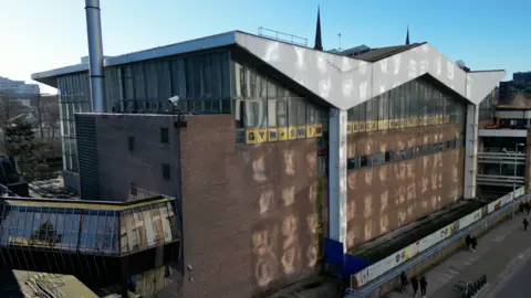 An aerial view of a multi-storey leisure centre complex, with windows near the top of a building on two visible sides of it. Lettering on the outside includes the words gym, swim and dance.
