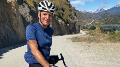 David is on a road with his bike wearing cycling attire. He is also wearing a helmet and smiling at the camera. Mountains can be seen in the distance.