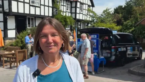 A woman with shoulder length brown hair, wearing silver hooped earrings and a silver necklace is smiling at the camera. She is wearing a blue top and a white cardigan. Behind her is a sign written people carrier with its boot open. It is parked on a road in front of a tudor style balck and white building.
