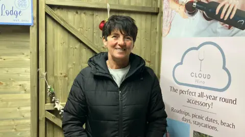 Denise McGilligan stands in front of a wooden door, with a sign promoting her wine business to her left. She has short dark hair and is wearing a heavy black winter coat, zipped up over a grey jumper.