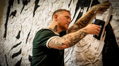 St David’s | Alistair Heap A close up of the mural. Wyburn has one arm against the mural and in his other hand he has a staple gun. He is wearing a green t-shirt with white trim, has short brown hair, a scruffy reddish beard and is heavily tattooed on both arms with and large monochrome butterfly tat on the right side of his neck.