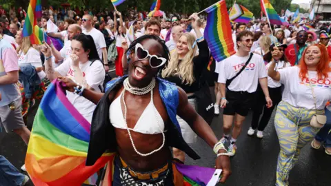 Pride revellers take part in the annual parade in London in 2025. Some people are waving rainbow flags and wearing face paint. 