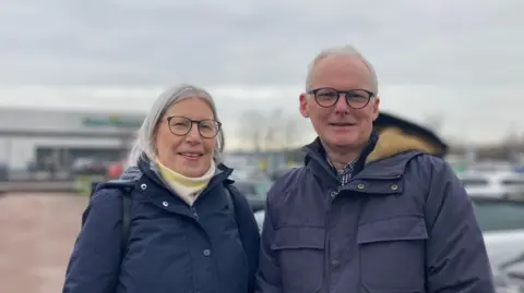 Jayne and Barrie are smiling, both are wearing blue warm winter coats. Jayne who has bob style grey hair and wears teal metal rimmed glasses, also wears a leather style rucksack, the straps are visible and a white and pale yellow neck scarf.
Barrie wears round dark rimmed glasses and a checked shirt just visible at the neck of his coat. The background is slightly blurred but you can just make out some cars and Morrisons signage in the distance.