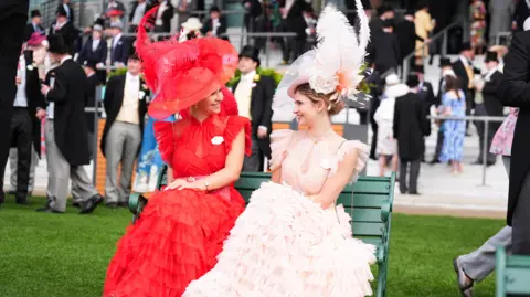 PA Two women in red and peach dresses and hats on a bench on the first day of Royal Ascot.