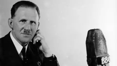 A black and white image of a man wearing a dark suit and tie with short neat hair and small moustache, looking straight at the camera with his head resting in his hand. Next to him is an old fashioned microphone with 'BBC' written on