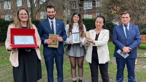 A school head teacher stands alongside four relatives of Wacław Brzeziński. They are holding memorabilia which they intend to swap 