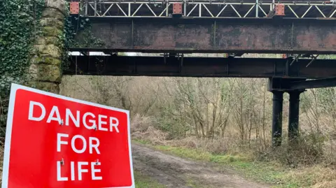 A red sign saying 'Danger for life' in front of a large bridge with a footpath beneath it. The bridge is in a rural area with lots of trees. 