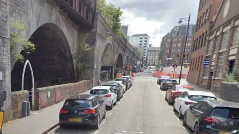 A google maps street view of a road with cars parked on both sides. A bridge with arches runs along the left hand side of the photo, with tall office buildings on the other side
