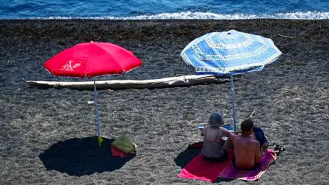 A red and blue parasol on a beach next to the sea, which is at the top of the picture. Two beachgoers are resting in the shade under the blue parasol on the right.