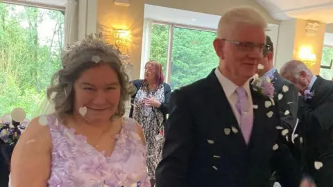 A close-up of Tina and John walking on their wedding day. She is dressed in a wedding dress and he is wearing a white shirt, pink tie, and a dark jacket. They are both smiling and walking under confetti.