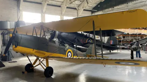 Bottisham Airfield Museum Tiger Moth N6635 in a hangar. It has a grey body with a bright yellow underbelly and two sets of bright yellow wings. Other aeroplanes can be seen behind it.