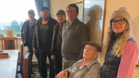 Local Democracy Reporting Service Five older men - including one wearing a flat cap, a chequered shirt and braces who is sat down - and one woman with long blonde hair, a beanie, and glasses stand in a pub.