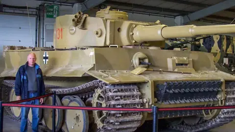 The Tank Museum Dale Oscroft, a who is bald and wearing a coat and blue jeans, pictured next to the Tiger 131, a brown/grey tank.