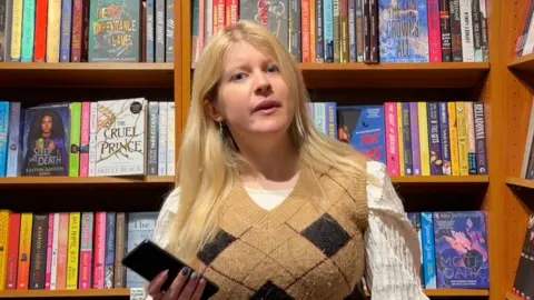 A young woman in a mustard checked top and white shirt, with blonde hair, in a book shop.