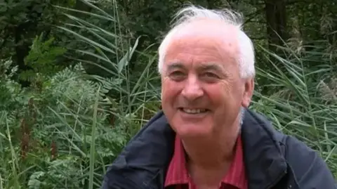 BBC Prof Alastair Fitter wearing a red shirt with white stripes and a blue waterproof coat photographed in front of bracken and reeds at Askham Bog