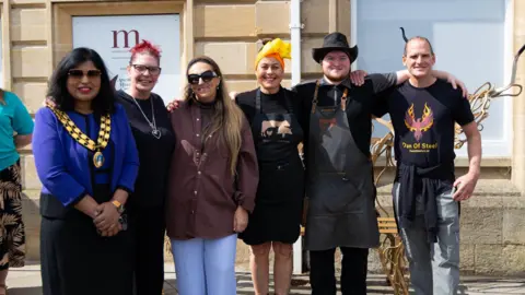Bertie Coy Six people stand in a row arm in arm and pose for a photo. The mayor is wearing chains and stands ont he far left. 