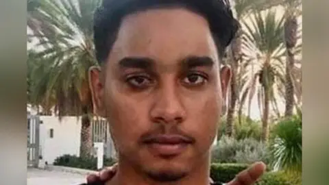 A close-up of a young man's face. He has dark hair and eyebrows and a sparse moustache and goatee beard. He is outside, surrounded by palm trees and low shrubs. A white wall and gates can be seen in the background.