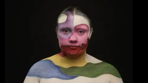 BBC/Wall To Wall/Dave King Ailish is looking directly into the camera with a completed make-up look. We can see her from her shoulders up and she has painted herself with a white base layer. On top of this are swirls of colour that have been painted on with brushtroke texture. One of her eyes is encircled with lilac. The other eye and middle part of her face is pink. Her lower lips and chin are covered in red. Her lower chin is orange and underneath this on her neck is yellow. One of her shoulders is blue while the other is green. Ailish's hair has been slicked back and she is against a plain black background. She has two silver nose pierecings in, one is a hoop and one is a stud.